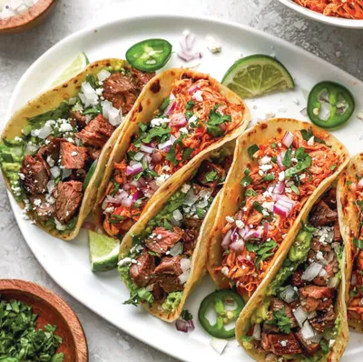 Row of loaded street tacos with grilled meat, guacamole, diced onion, cilantro, and jalapeños on a white platter