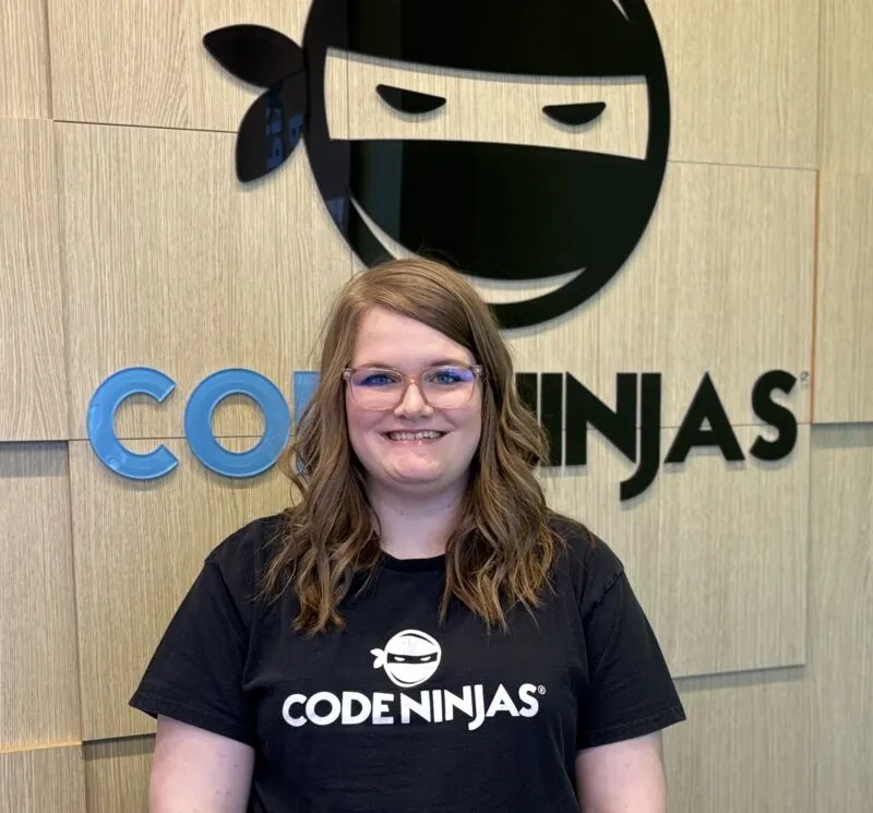 Smiling woman in a Code Ninjas T-shirt standing before the Code Ninjas logo on a wood-panel wall