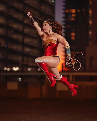 Woman in Wonder Woman cosplay leaps mid-air against a night city skyline