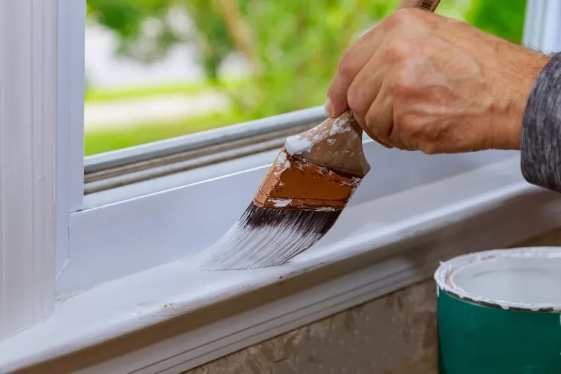 Hand applying white paint to a window sill with a wide brush, paint can nearby