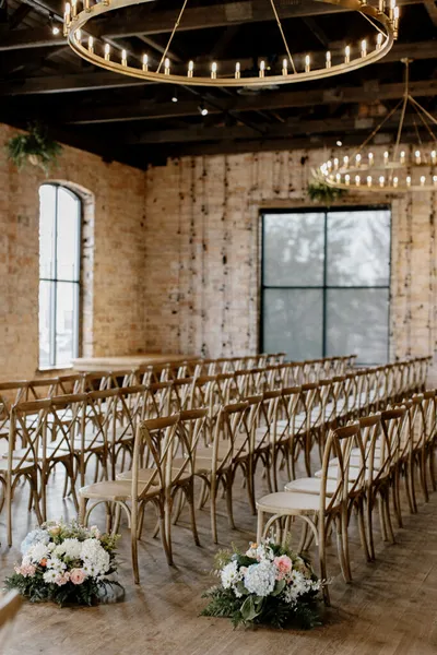 Rows of wooden cross-back chairs along a floral-lined aisle in a brick-walled venue with circular chandelier