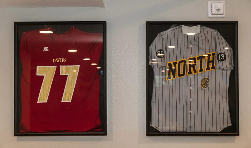 Two framed sports jerseys on a wall — a red football jersey numbered 77 and a gray pinstriped baseball jersey reading 'North