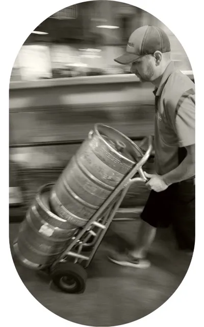 Worker in cap hauling stacked beer kegs on a hand truck, motion blur