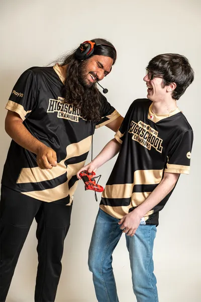 Two laughing players in black-and-gold High School Champions esports jerseys share a game controller