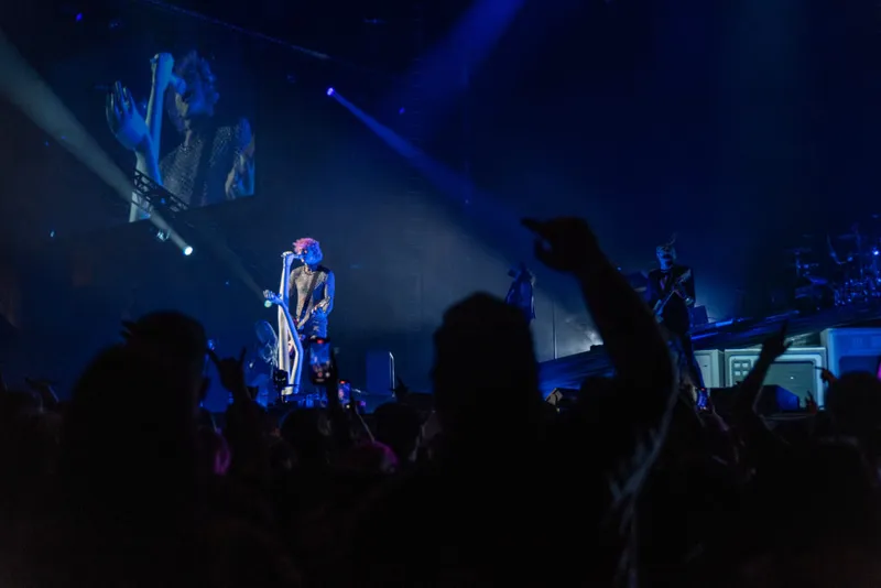 Performer with pink hair in a sequined outfit sings on a dark stage lit by blue beams above a packed crowd