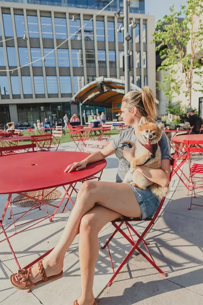 Woman in denim shorts holds a small Pomeranian at a red bistro table on a sunny urban patio