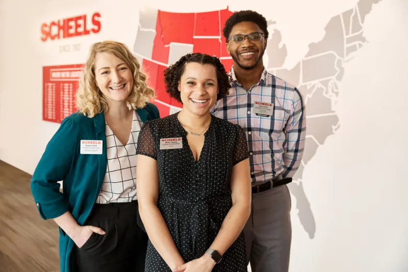 Three smiling Scheels employees wearing name badges stand before a US map mural