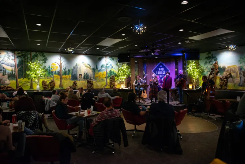 Live band performs on a small stage at the dimly lit Troll Lounge, with guests seated at tables amid forest-themed murals