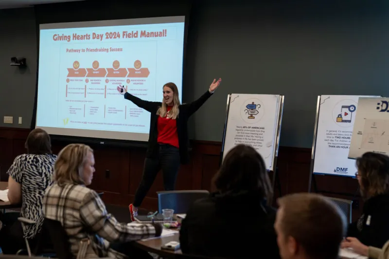 Woman presenting Giving Hearts Day 2024 Field Manual slideshow to seated attendees in a conference room