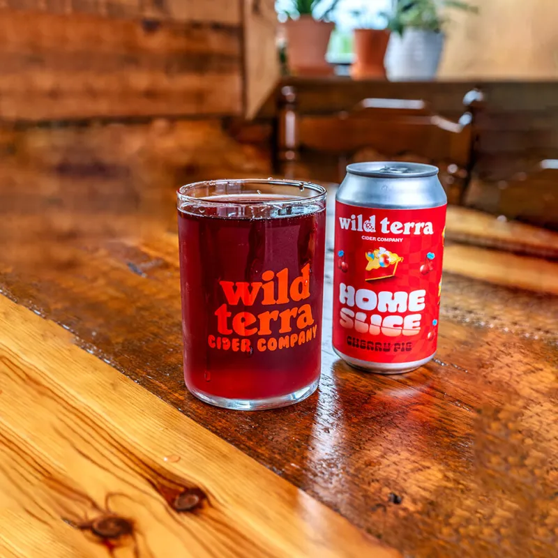 Wild Terra Cider Company glass of dark red cider beside a Home Slice cherry pie cider can on a wooden bar