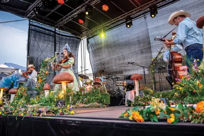 Band performs on a flower-decorated outdoor stage with giant mushroom props and string instruments