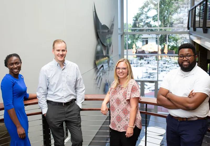 Four smiling professionals posed on an indoor mezzanine balcony near large windows