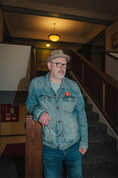 Man in denim jacket and fedora leaning on a staircase railing inside an older building