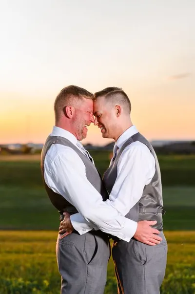 Two grooms in matching gray vests embrace forehead-to-forehead at sunset over an open field