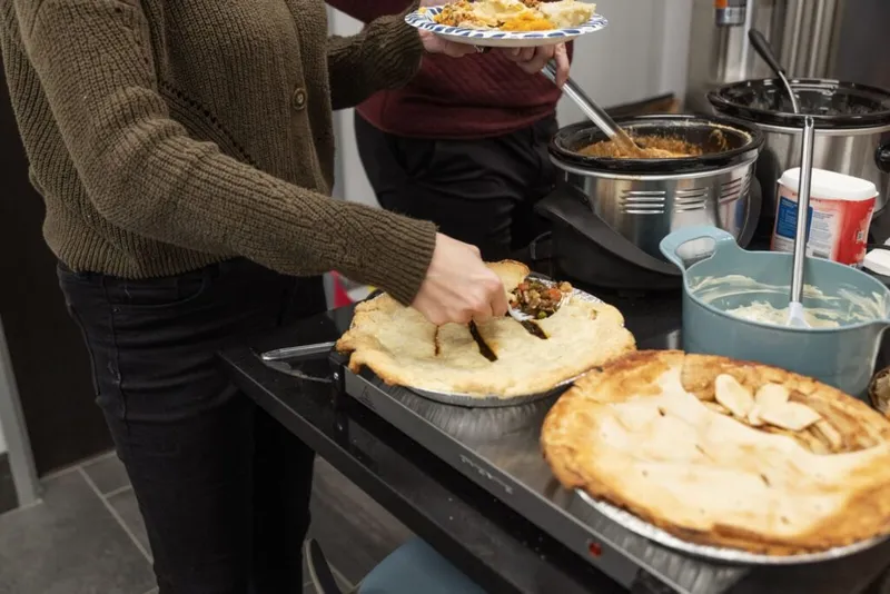 Two people serving food at a potluck spread with two golden pies and slow cookers
