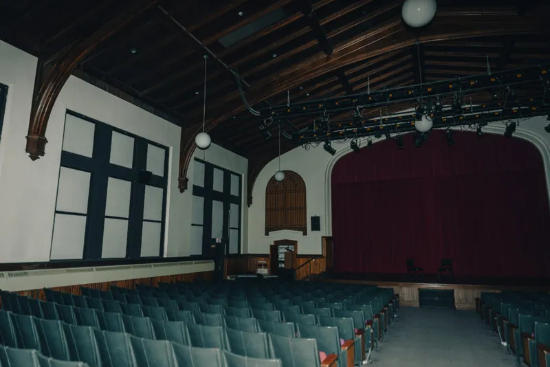 Empty auditorium with rows of blue seats, wood-beam ceiling, stage lights, and red curtain