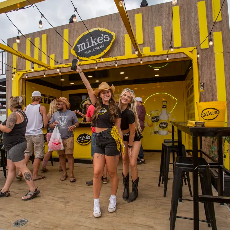 Two women pose for a selfie in front of a yellow Mike's Hard Lemonade festival booth