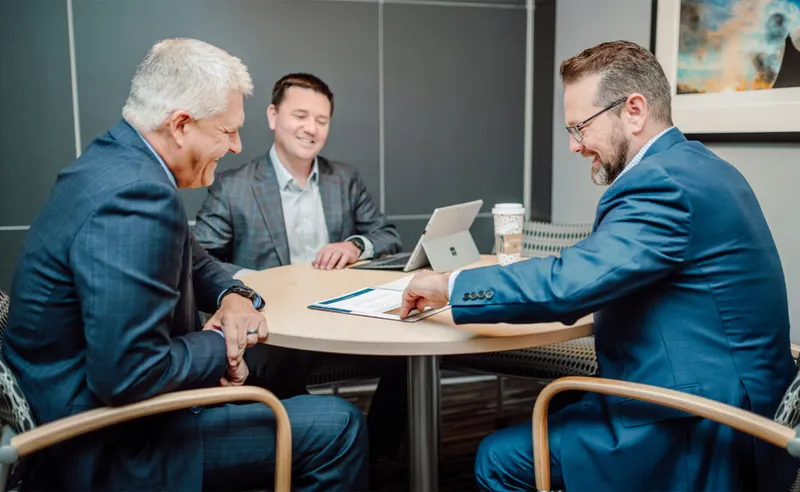 Three men in suits smiling around a small table with a tablet and documents