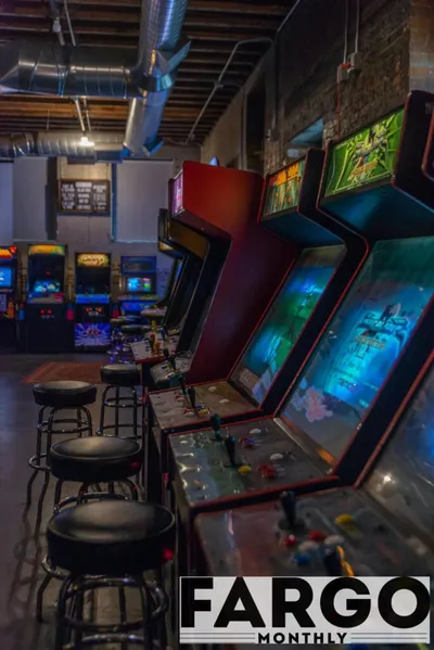 Row of vintage arcade cabinets with glowing screens and bar stools in a dimly lit brick-walled venue