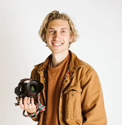Smiling young man with blond hair holding a DSLR camera, wearing a tan jacket
