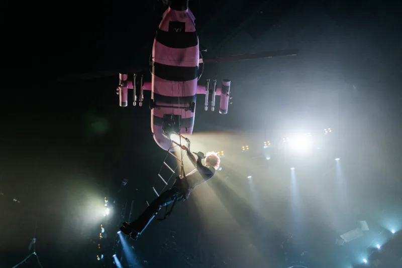 Performer dangling from a pink and black striped flying prop above a darkened arena stage