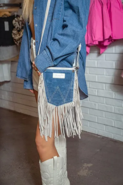 Woman in denim shirt and white cowboy boots wearing a fringe crossbody bag