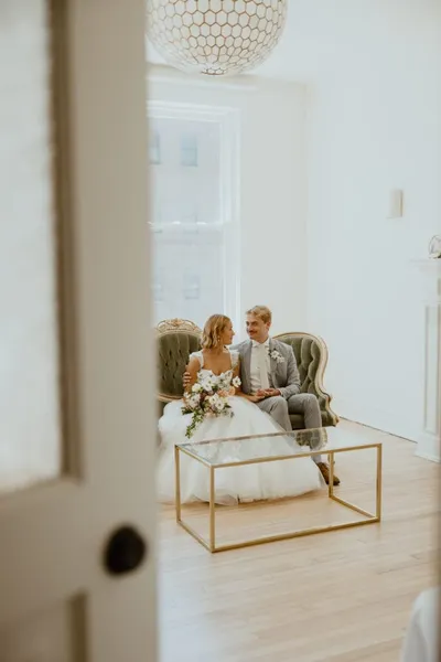 Bride and groom seated together on a green velvet chair, smiling at each other in a bright white room
