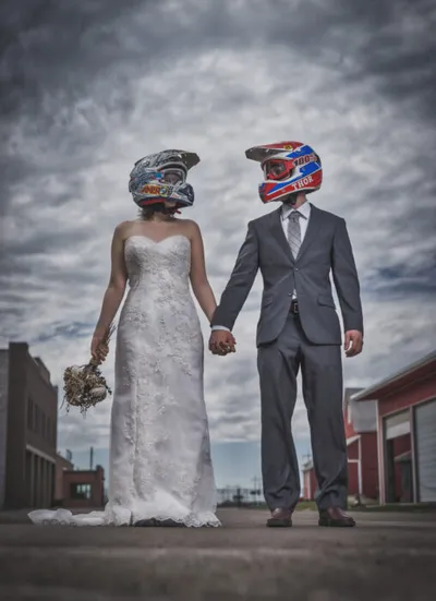 Bride in white gown and groom in gray suit holding hands while wearing motocross helmets under a cloudy sky