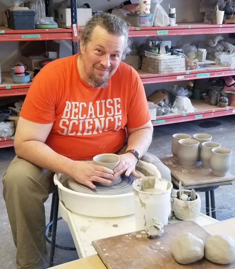 Bearded man in orange 'Because Science' shirt shapes clay on a pottery wheel in a cluttered studio