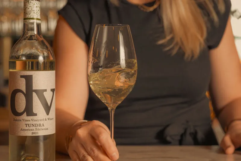 A glass of white wine beside a Dakota Vines Tundra 2021 bottle held by a woman in a black top