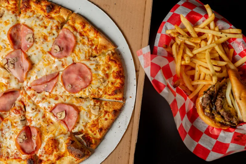 Ham and mushroom pizza in a box beside a burger and fries in red-checkered paper