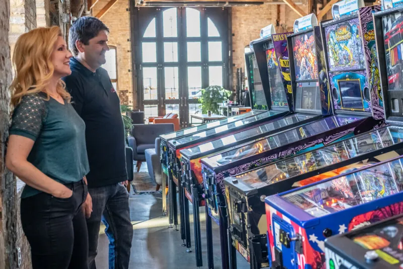 A woman and man smile while looking at a row of pinball machines in a brick-walled venue