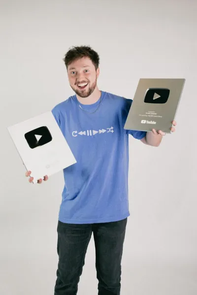 Smiling bearded man in blue T-shirt holding two YouTube Play Button awards