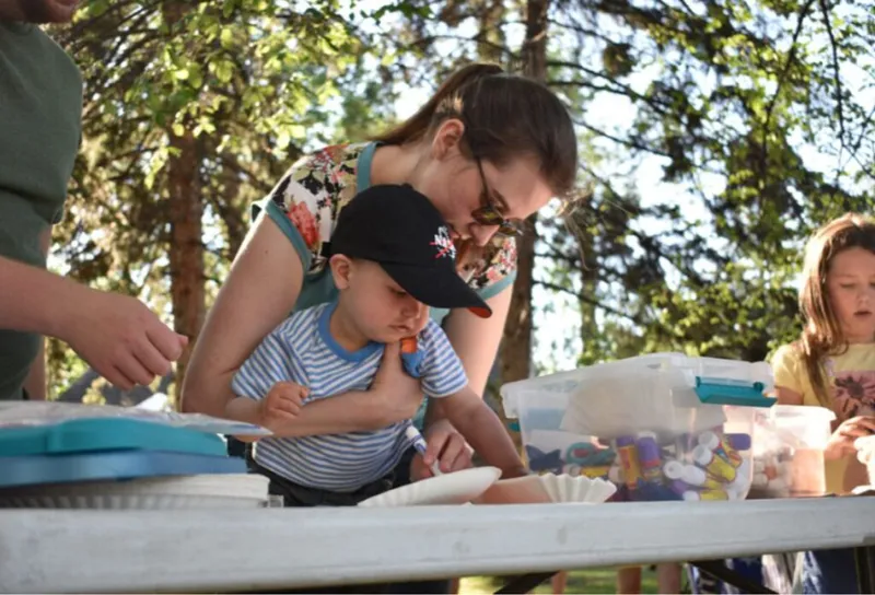 Woman helping a young child with a craft project at an outdoor picnic table