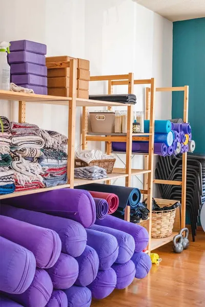 Wooden shelves stocked with rolled yoga mats, folded blankets, purple foam blocks, and bolsters in a studio supply room