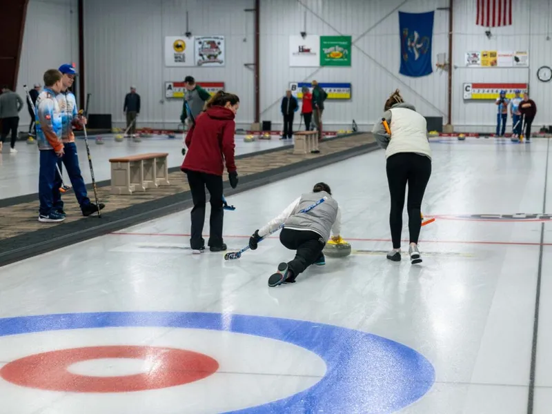 Curlers on an indoor ice sheet, one player crouched releasing a stone toward the target rings