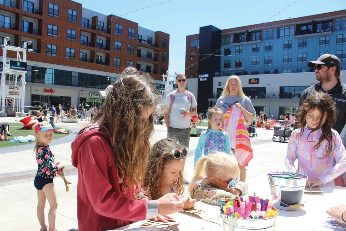 Children doing crafts at an outdoor table in a busy urban plaza on a sunny day